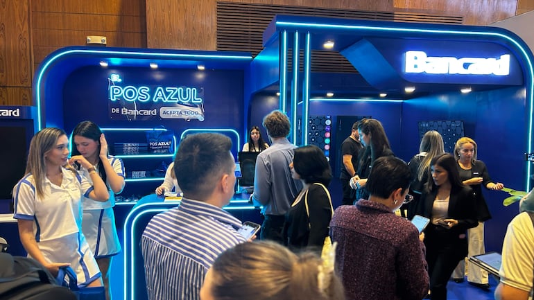 Mujeres en uniformes blancos y azules en el stand de Bancard, conversando y acomodándose en un ambiente iluminado con luces LED.