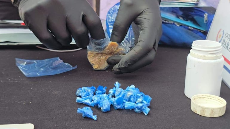 Una mano con guantes negros sostiene un paquete, rodeado de envoltorios azules y un recipiente blanco en una mesa oscura.