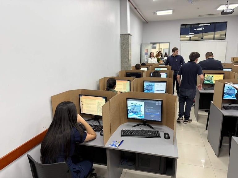 Tres estudiantes se enfocan en computadoras, mientras en el fondo tres personas conversan en un aula blanca y brillante.