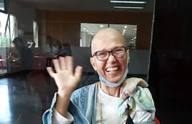 Norma Beatriz, con gafas y camiseta blanca, sonríe y saluda, rodeada de un ambiente acogedor y familiar.