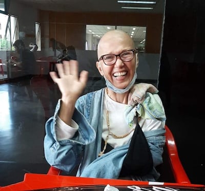 Norma Beatriz, con gafas y camiseta blanca, sonríe y saluda, rodeada de un ambiente acogedor y familiar.