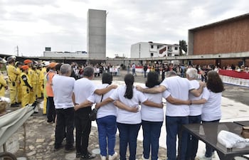 Multitud de personas con camisetas blancas abrazándose en un evento conmemorativo, rodeados de edificios urbanos y miembros de un equipo de rescate.