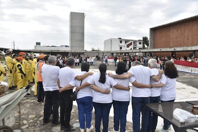 Multitud de personas con camisetas blancas abrazándose en un evento conmemorativo, rodeados de edificios urbanos y miembros de un equipo de rescate.