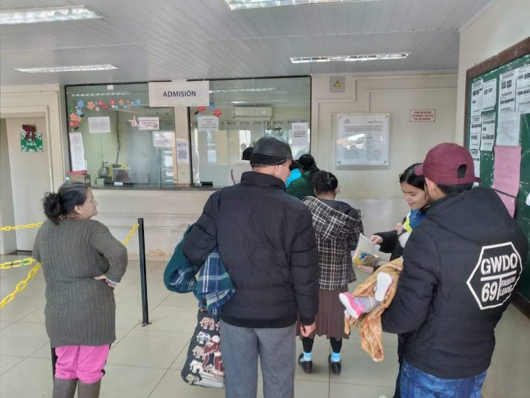 Mujer de pie en perfil y tres personas más conversan en una oficina de admisión. Paredes claras y ventanilla visible.