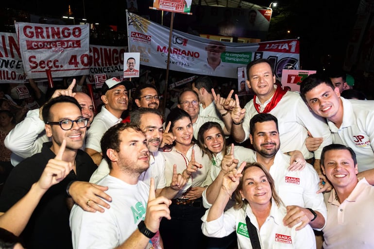 Miguel Sosa y Costanza Troche, sonriendo y mostrando gestos de triunfo, rodeados de personas en camisetas blancas con pancartas políticas.