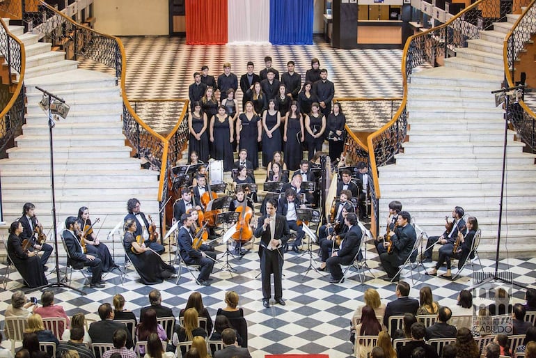 Músicos de orquesta y coro en el escenario, vestidos de negro, dirigiendo un evento decorado con los colores nacionales.