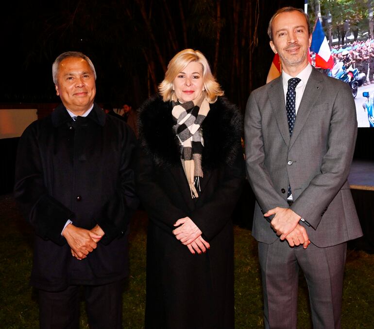Mujer rubia en abrigo negro y bufanda a cuadros, hombre canoso con abrigo oscuro y hombre de barba con traje gris, todos sonriendo.