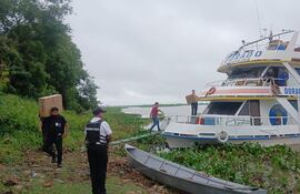 Hombres en la orilla del río, uno lleva una caja sobre su hombro, un barco anclado con el nombre 'El Dorado' al fondo.