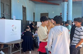Grupo de jóvenes discutiendo en un ambiente electoral, rodeados de mesas y una urna de votación con el emblema de Paraguay.