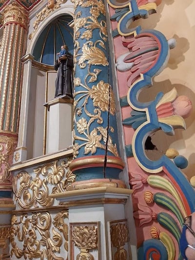 Interior de un templo religioso adornado con detalles ornamentales en tonos azul, rosa y dorado, con una estatuilla de un santo a la izquierda.