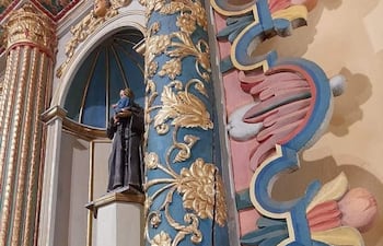 Interior de un templo religioso adornado con detalles ornamentales en tonos azul, rosa y dorado, con una estatuilla de un santo a la izquierda.