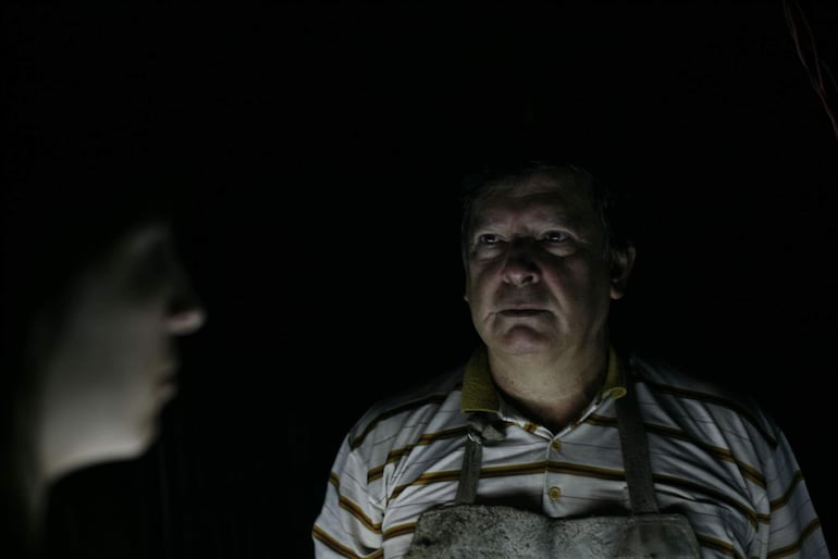 Hombre de mediana edad con camisa a rayas y delantal, observa a una mujer de cabello largo en un entorno oscuro y serio.