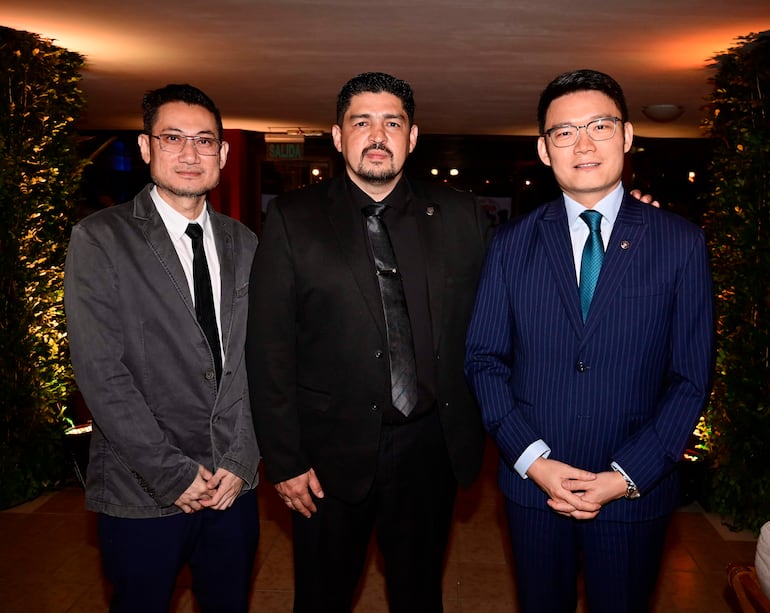 Tres hombres sonrientes en el evento, uno en chaqueta gris, otro en traje negro y el tercero en traje a rayas azul.