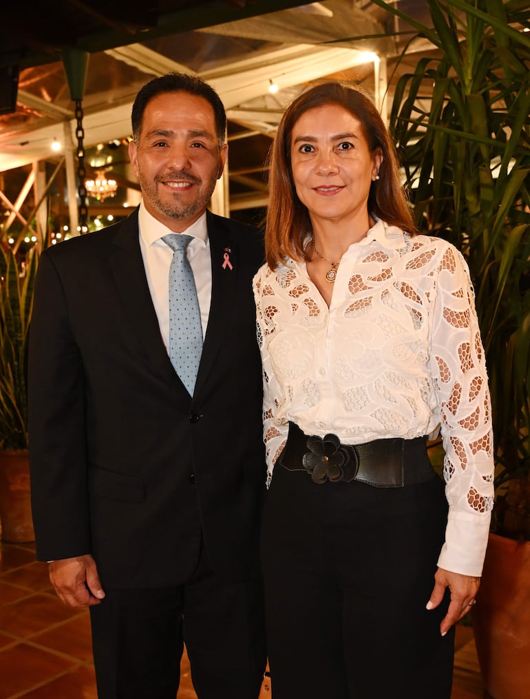 Hombre con traje oscuro y corbata celeste y mujer en blusa blanca sonríen en un ambiente elegante con plantas y luces suaves.