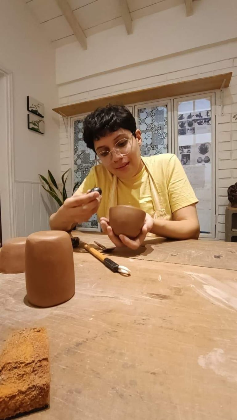 Joven con cabello corto y gafas, viste camiseta amarilla, trabaja con barro en un taller de cerámica con paredes blancas.
