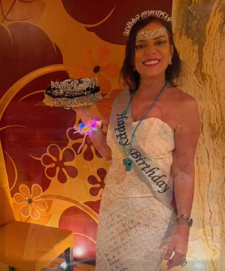 Mujer sonriente en vestido blanco de encaje con diadema, sostiene un pastel negro y un objeto colorido en fiesta de cumpleaños.