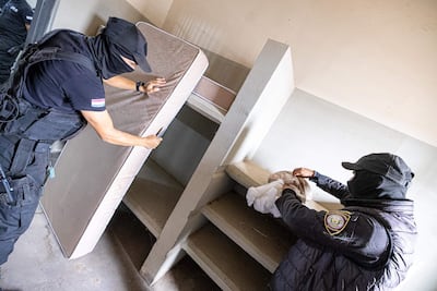 Dos agentes de seguridad con pasamontañas inspeccionan colchones en un ambiente cerrado y austero.