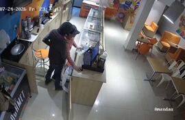 Hombre de gorra y ropa oscura detrás del mostrador mientras cajera muestra gesto de tensión en heladería colorida.