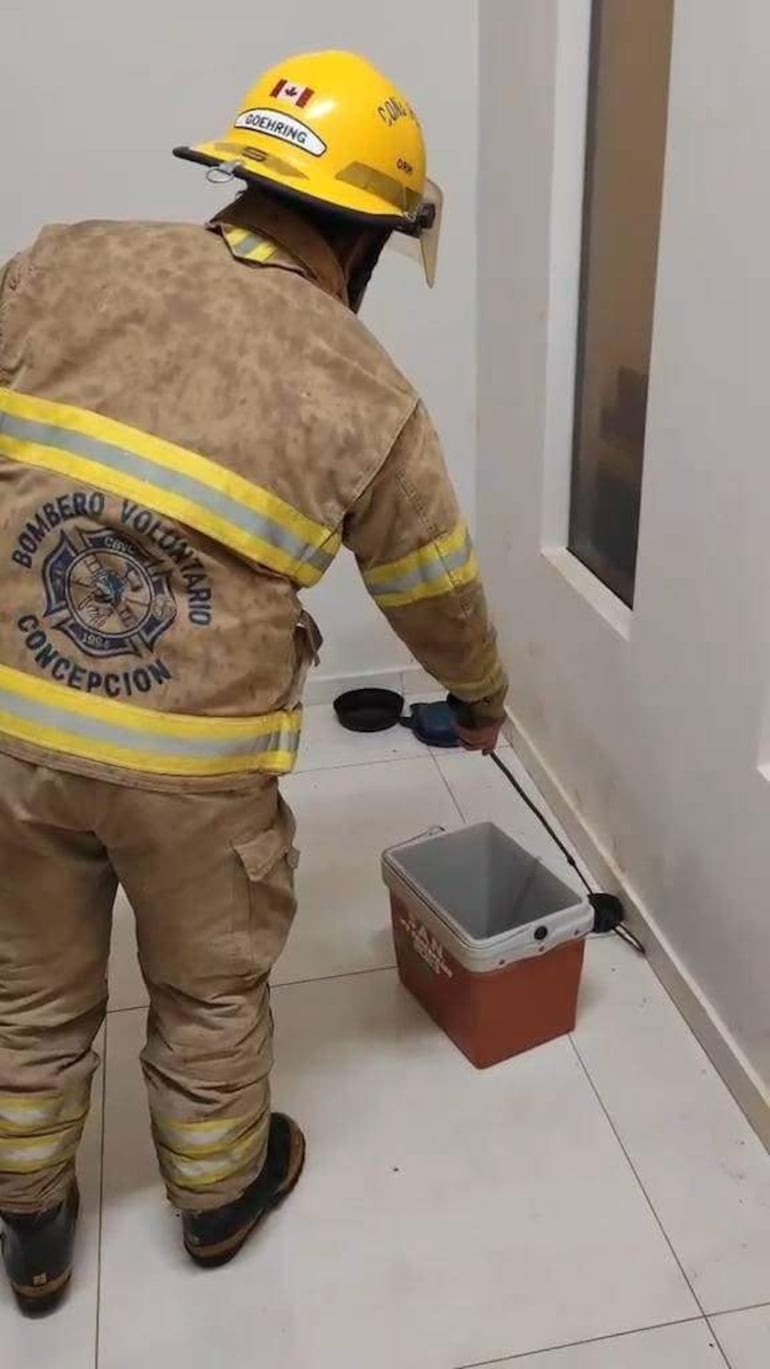 Bombero voluntario con casco amarillo y uniforme, agachado frente a un recipiente marrón en un ambiente limpio y ordenado.