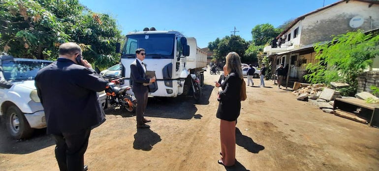 Dos hombres conversan junto a un camión blanco mientras una mujer escucha de espaldas. Ambiente urbano con vegetación.