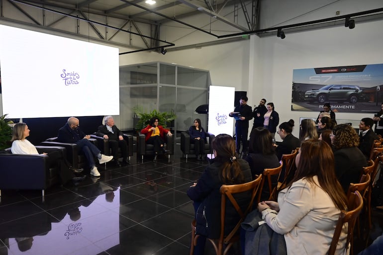 Cinco personas sentadas en un panel, una mujer de cabello largo y otra con abrigo rojo, ambiente formal frente a pantallas con texto sobre el evento.