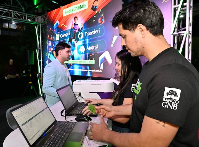 Hombre en blazer claro dialoga, mientras dos personas en camisetas de Banco GNB atienden públicos con laptops sobre mesa.