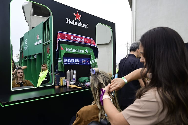 Mujer rubia recibe tratamiento de peinado mientras se ven bufandas del Arsenal y PSG, con logo de Heineken en fondo verde.