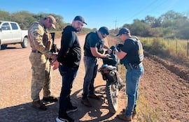 Hombre de espaldas siendo registrado por un agente armado en uniforme camuflado, otros agentes examinan una mochila en el campo.