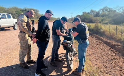 Hombre de espaldas siendo registrado por un agente armado en uniforme camuflado, otros agentes examinan una mochila en el campo.