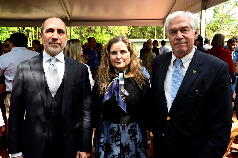 Tres personas en primer plano: hombre de traje oscuro y corbata clara, mujer con blusa negra y falda floral, hombre canoso con traje y corbata azul claro en un evento al aire libre.