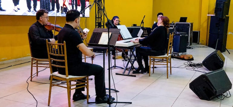 Cinco músicos, entre ellos una mujer en teclado y dos hombres con instrumentos, se presentan en un escenario con paredes amarillas.