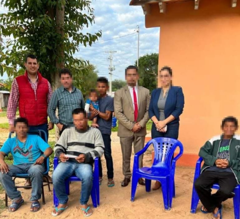 Grupo de seis personas en entorno rural; varios hombres sentados, con camisetas de colores y una vivienda naranja de fondo.