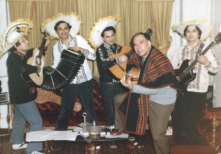 Cinco músicos con sombreros decorados y camisas coloridas tocan guitarra y acordeón en ambiente acogedor con sofá y cortinas.