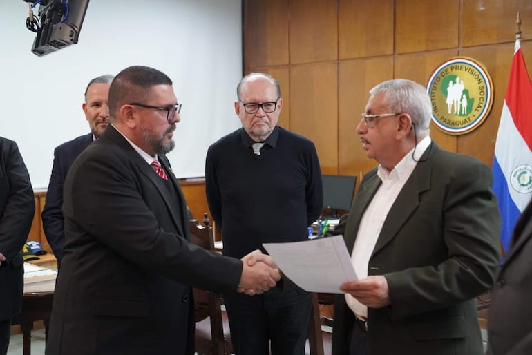 Isaías Fretes con saco negro y corbata roja estrecha la mano de Hassel Jiménez en oficina del IPS, rodeados de documentos.