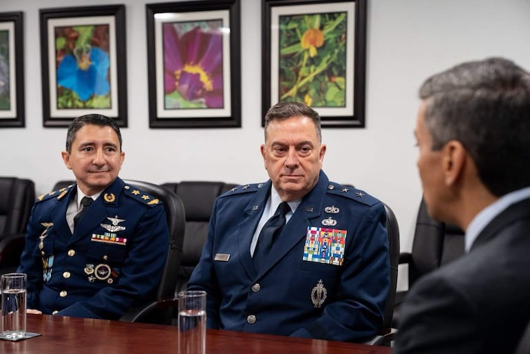 Santiago Peña en traje oscuro habla con dos oficiales militares en uniforme, en un entorno de oficina formal y decoraciones florales.