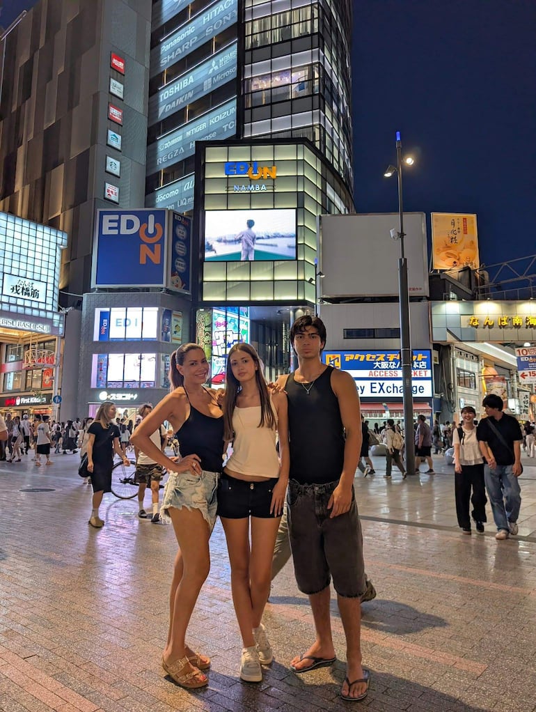 Dos mujeres en camiseta y shorts, y un hombre en camiseta negra, sonríen en una concurrida calle con anuncios de neón.