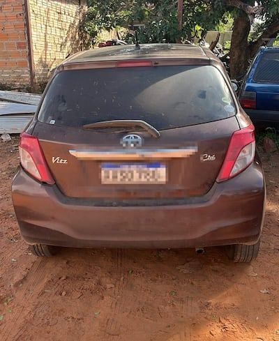 Vehículo Toyota Vitz color marrón estacionado en una superficie de tierra, con motos y otro auto al fondo.