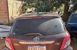 Vehículo Toyota Vitz color marrón estacionado en una superficie de tierra, con motos y otro auto al fondo.