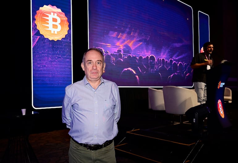 Hombre de pie frente a pantalla grande sobre Bitcoin, con expresión tranquila, en un ambiente interior de salón de eventos.