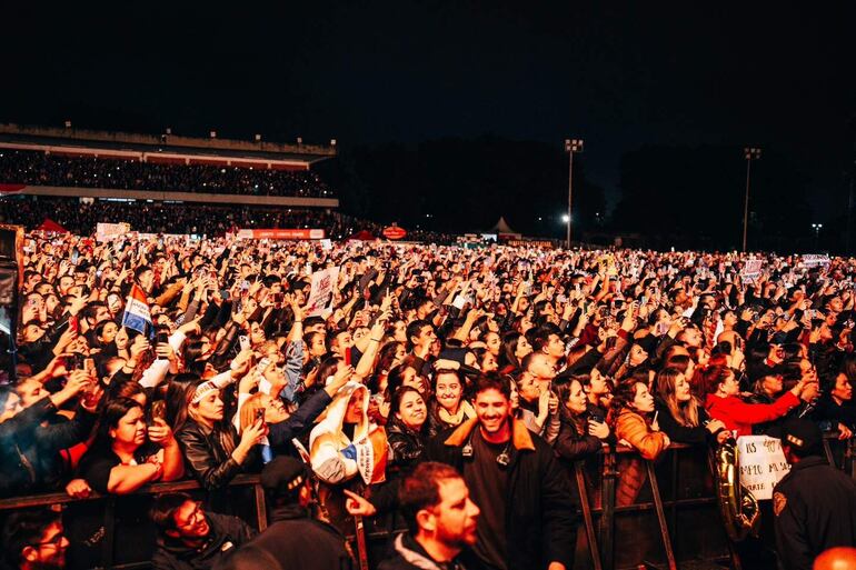 Multitud de jóvenes sonrientes en concierto, sosteniendo teléfonos y pancartas, creando una atmósfera vibrante y festiva.