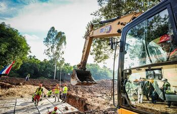 Seis trabajadores en chalecos amarillos y cascos, algunos en el suelo y uno en excavadora CAT amarilla, en un entorno vegetal.