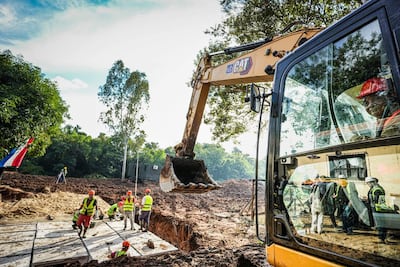 Seis trabajadores en chalecos amarillos y cascos, algunos en el suelo y uno en excavadora CAT amarilla, en un entorno vegetal.