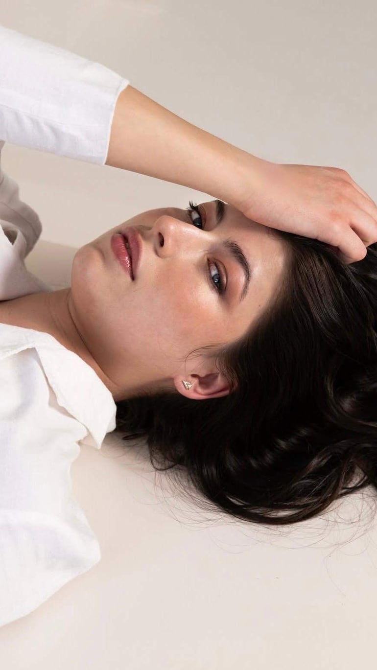Joven mujer de cabello oscuro y ondulado, viste blusa blanca, posando sobre una superficie clara con una expresión serena.