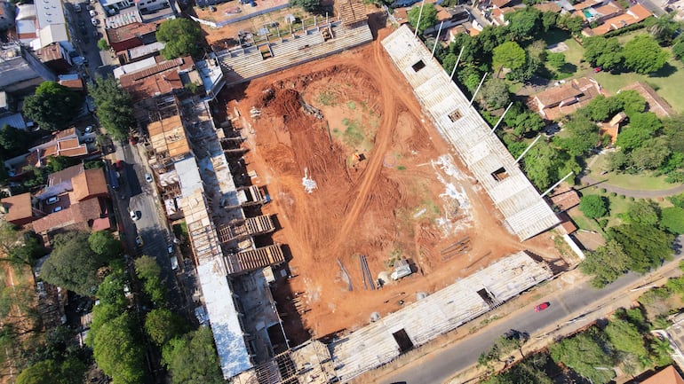 Vista aérea de obra en construcción con estructuras metálicas y maderas, rodeada de casas y árboles.