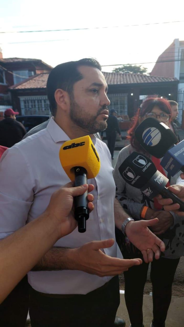 Luis Bello, con camisa blanca y barba, gesticula mientras habla a un grupo de periodistas con varios micrófonos.
