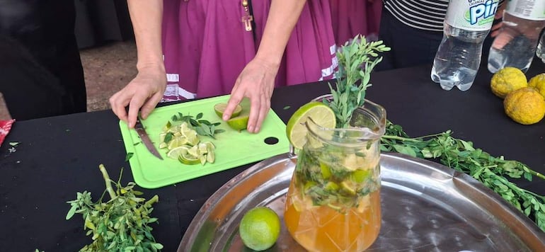 Cocinera con blusa fucsia pica limones en tabla verde, con jarra de bebida y frutas de fondo.