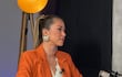 Mujer sentada en sofá con traje naranja, cabello en coleta alta y grandes aretes, en conversación en un estudio.