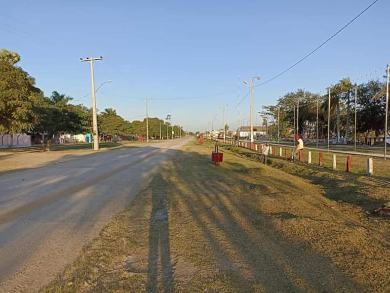 Dos trabajadores cerca de postes de luz, una persona inclinada cuidando el césped en una calle desierta rodeada de árboles.