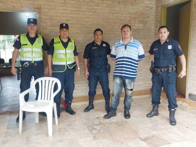 Hombre de camiseta a rayas rodeado de policías en uniformes, en ambiente con paredes de ladrillo y silla plástica vacía.