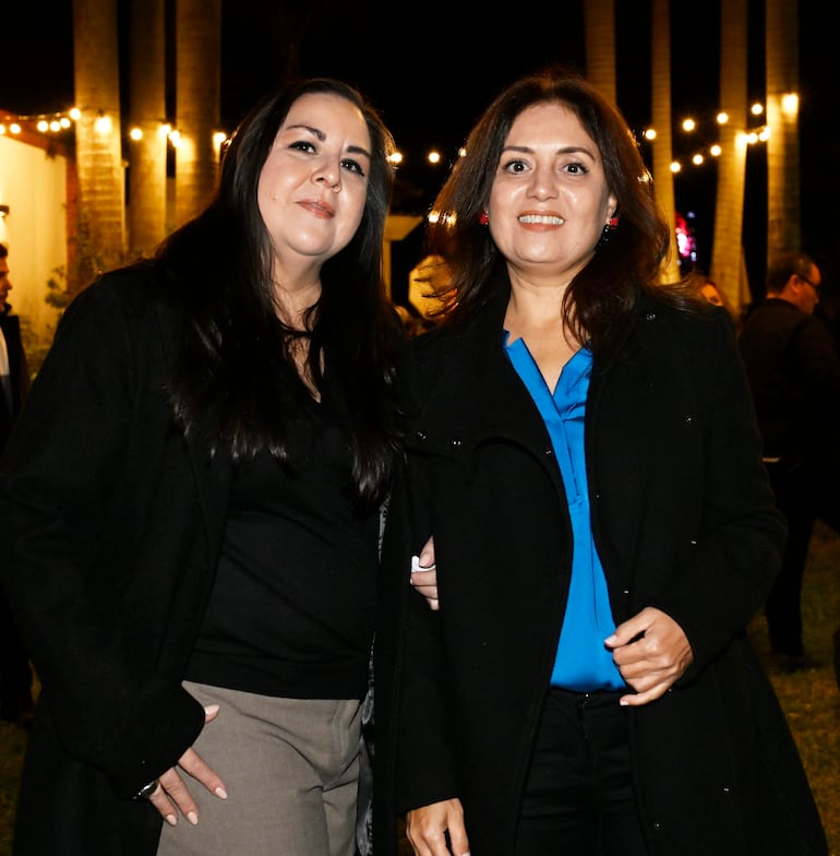 Dos mujeres sonrientes en abrigos oscuros, posando en un ambiente festivo con luces colgantes en un espacio exterior.